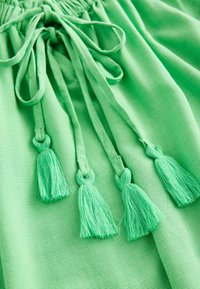 Indumento verde caratterizzato da un tessuto testurizzato e una chiusura con cordoncino e nappine. Le nappine sono anch'esse di una tonalità verde coordinata.