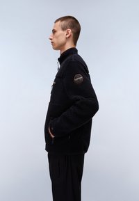 Schwarze Fleecejacke mit strukturiertem Finish, ausgestattet mit einem Reißverschluss, seitlichen Taschen und einem kontrastierenden Stoffeinsatz auf der Brust.