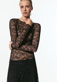 Črna majica z dolgimi rokavi iz čipke z cvetličnim vzorcem in prosojno teksturo, nošena s pripadajočo črno čipkasto krilom. Modelka pozira z zloženimi rokami.
