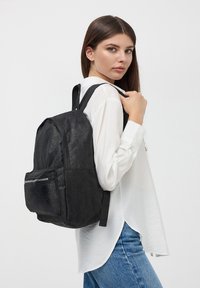 Zaino in tessuto nero con superficie texture, dotato di tasca frontale con zip e spallacci regolabili, indossato da un modello con una camicia bianca.