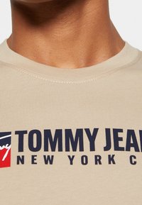 Beige short-sleeve T-shirt with a ribbed neckline, featuring bold navy text reading "TOMMY JEANS NEW YORK" and a small color logo accent.
