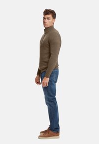 Pulover zip-up maro cu manșete cu nervuri, asortat cu blugi albaștri și adidași din suede maro. Puloverul are un mic logo pe piept.