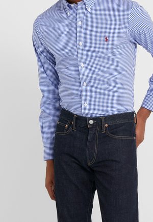 Man draagt een blauw-wit geruit overhemd met knopen in een donkerblauwe spijkerbroek, met de hand op de heup.