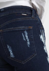 Vaqueros de mezclilla azul oscuro con detalles desgastados, con desgarros en varias partes, costuras en contraste y un diseño de cintura media.