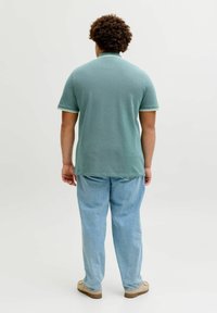 Άνδρας με σγουρά μαλλιά, φοράει πράσινο polo με κοντά μανίκια, ανοιχτό μπλε τζιν και μπεζ παπούτσια, στέκεται με την πλάτη στραμμένη σε ένα απλό φόντο.