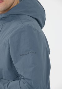 Ljusblå vattentät jacka med huva, med en texturerad tyg och en svart logotyp på vänster arm.