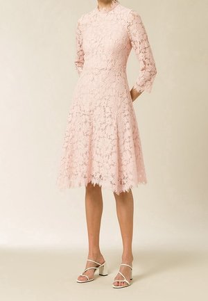 Robe en dentelle rose avec des manches trois-quarts, un corsage ajusté, une jupe évasée, un ourlet festonné et un motif floral. Associée à des sandales à talons blocs blanches.