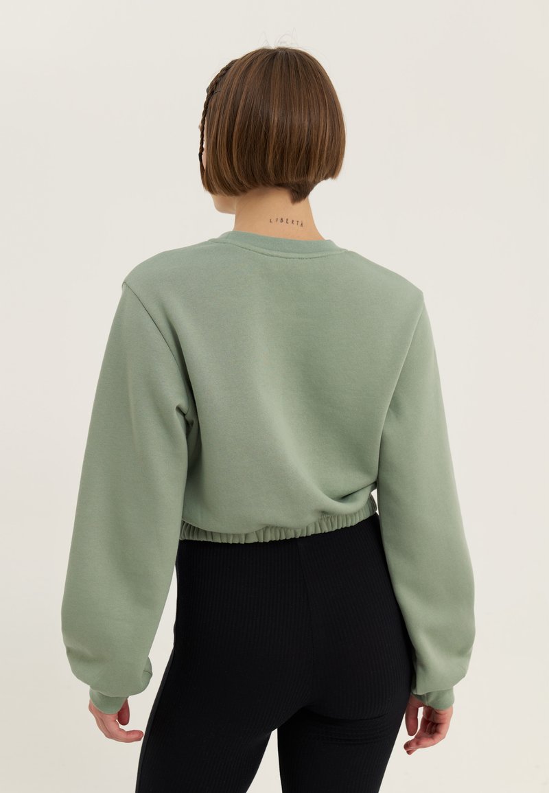 Sudadera verde corta con mangas largas, dobladillo acanalado y textura suave. Combinada con leggings negros acanalados, que presentan un diseño simple.