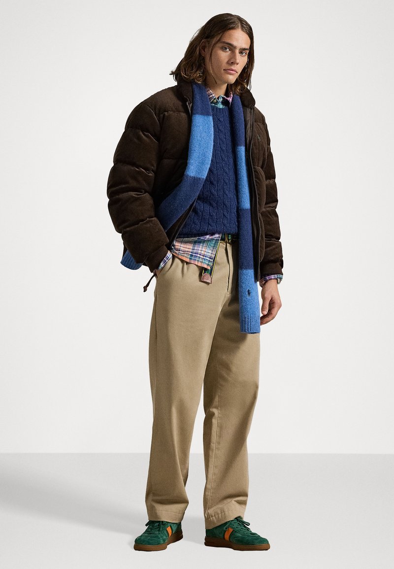 Doudoune marron, pull en tricot bleu à mailles, chemise à carreaux, pantalon kaki et baskets vertes avec des accents orange. Écharpe en tricot texturé de couleur bleue.