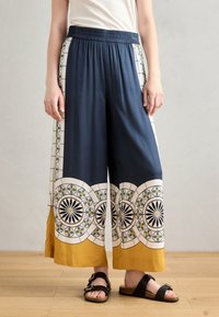 Navy wide-leg trousers with patterned white and mustard accents, featuring geometric designs and an elastic waistband. Worn with black sandals.