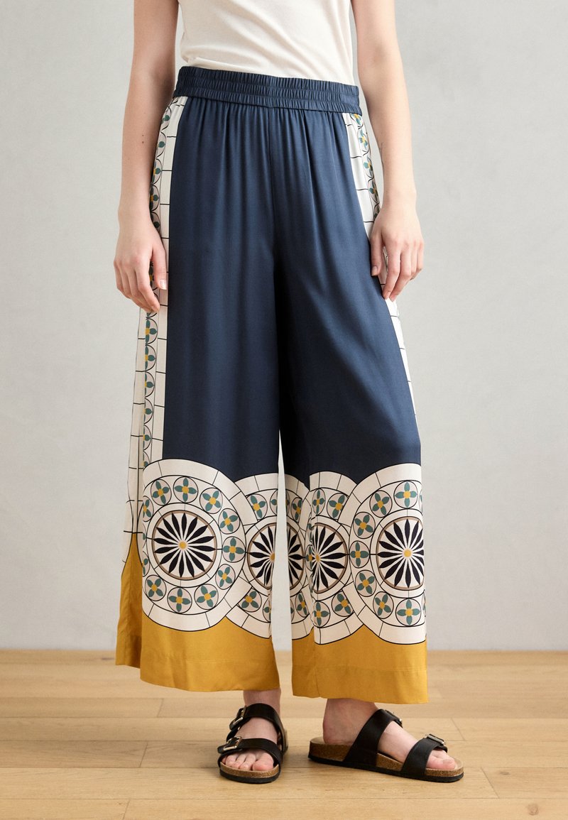 Navy wide-leg trousers with patterned white and mustard accents, featuring geometric designs and an elastic waistband. Worn with black sandals.