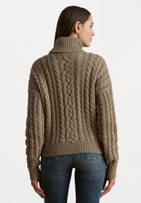 Chunky beige strikkegenser med høy krage, med kabelstrikk mønster og ribbestrikkede mansjetter og nederkant. Brukt over mørke jeans.