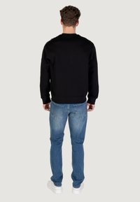 Hombre de espaldas vistiendo un sudadera negra, jeans azules con bolsillos traseros y zapatillas blancas, apoyado contra un fondo claro y liso.