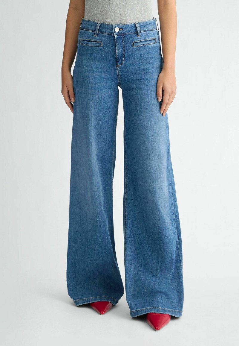 Jean en denim bleu à jambes larges avec poches avant, taille haute et texture lisse, associés à des talons aiguilles rouges.