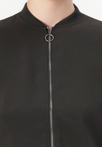 Närbild av en svart jacka med en silvrig dragkedja med ett runt draghandtag, som bärs av en person med ljus hudton.