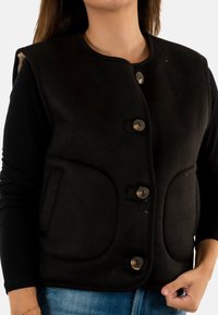 Gilet nero con scollatura tonda, bottoni decorativi, tasche laterali e una texture morbida. Indossato sopra una maglietta a maniche lunghe e jeans.