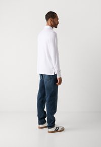Mann, der seitlich steht und ein weißes Langarmhemd, blaue Jeans und weiße Sneakers mit schwarzen Streifen auf einem einfarbigen Hintergrund trägt.