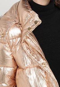 Detailní záběr na metallic růžově zlatou péřovou bundu se zipem a cvoky, kterou nosí přes černý rolák.