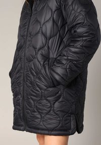 Veste rembourrée matelassée noire avec un motif rond, dotée d'une fermeture éclair sur le devant et de poches latérales. Tissu extérieur lisse et brillant avec des poignets côtelés.