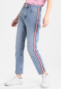 Vaqueros de corte recto, de cintura alta, en azul claro, con franjas laterales rojas y blancas, y un diseño clásico de cinco bolsillos, combinados con zapatillas blancas.