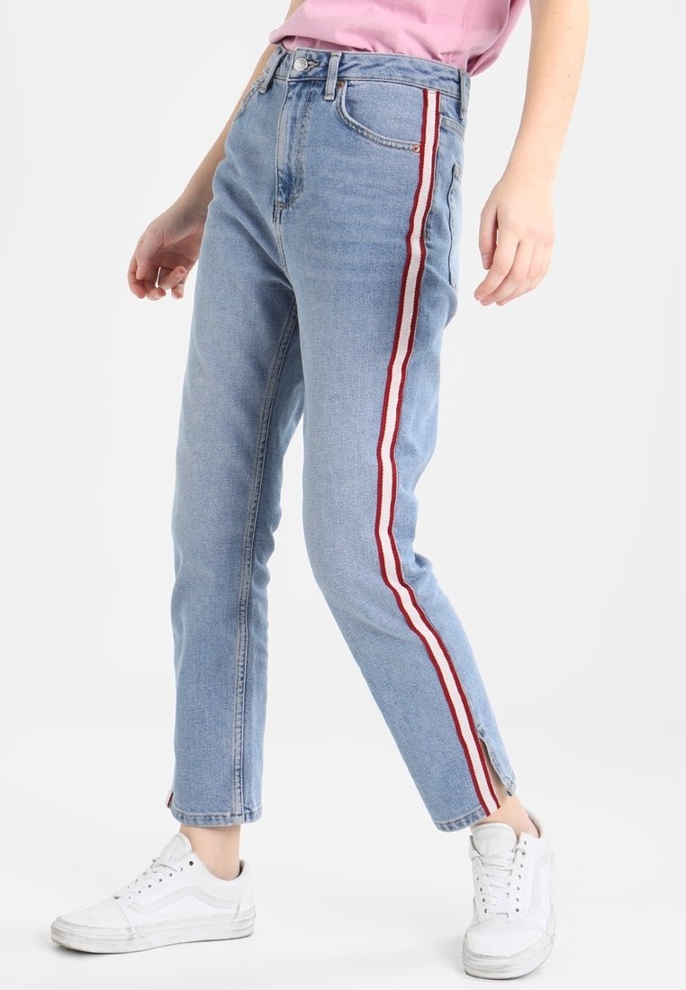 Vaqueros de corte recto, de cintura alta, en azul claro, con franjas laterales rojas y blancas, y un diseño clásico de cinco bolsillos, combinados con zapatillas blancas.