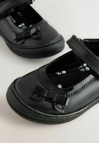 Schwarze Lederschuhe mit einer abgerundeten Zehe, einem Riemen über dem Spann und einem kleinen Schleifenakzent; strukturierte Gummisohle und gepolsterter Innenraum.