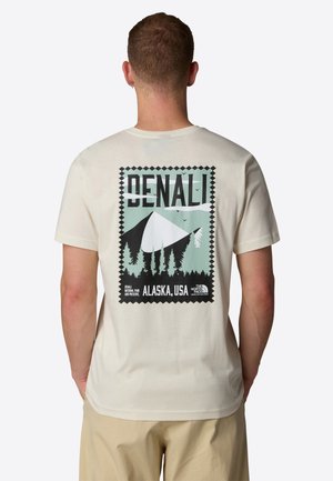 Mężczyzna w kremowej koszulce z grafiką przedstawiającą górę Denali i las oraz napisem "Denali Alaska, USA" na plecach.