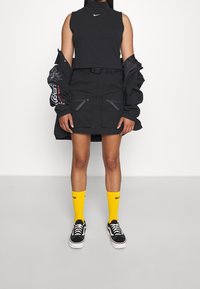 Svart crop top, svart kjol med dragkedjefickor, stor svart jacka med grafisk ärm, gula Nike-strumpor, svarta Vans-skor.