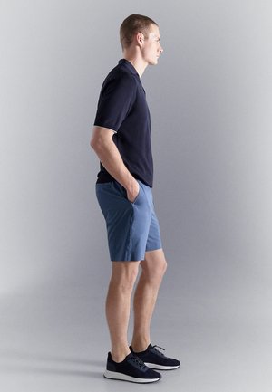 Jeune homme debout de profil, portant un polo noir, un short bleu et des baskets noires, les mains dans les poches, sur un fond uni.