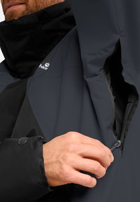 Veste imperméable noire et gris foncé, dotée d'un col haut, d'une poche zippée et de poignets ajustables avec fermetures velcro.