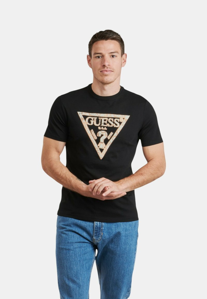 Camiseta de algodón negra con un triángulo dorado central que presenta el texto "GUESS?" y un signo de interrogación, combinada con jeans azules.