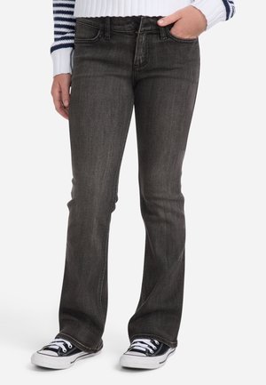 Person iført mørkegrå jeans med lige ben, sorte og hvide sneakers og en hvid sweater med marineblå striber, stående med den ene hånd i lommen.
