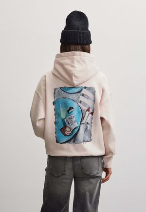 Person iført en lysebeige hættetrøje med farverigt grafik af bord, drik, bog, sko og kamera på bagsiden, kombineret med mørke jeans og sort hue.
