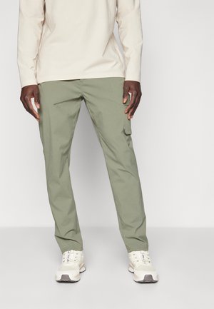 Mand iført lysebeige langærmet skjorte, olivengrønne cargobukser med sidelomme og hvide sneakers, stående mod en ensfarvet baggrund.