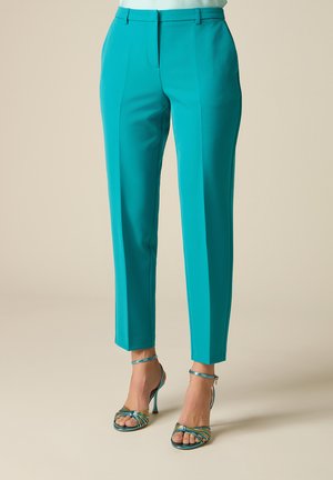 Teal op maat gemaakte enkelhoge broek gedragen met multicolor sandalen met hoge hak en bandjes op een neutrale achtergrond.