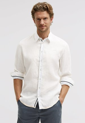 Mann mit kurzen, lockigen Haaren, der ein weißes, langärmliges Hemd mit aufgerollten Ärmeln und dunkle Hosen trägt, Hände in den Taschen, neutraler Hintergrund.