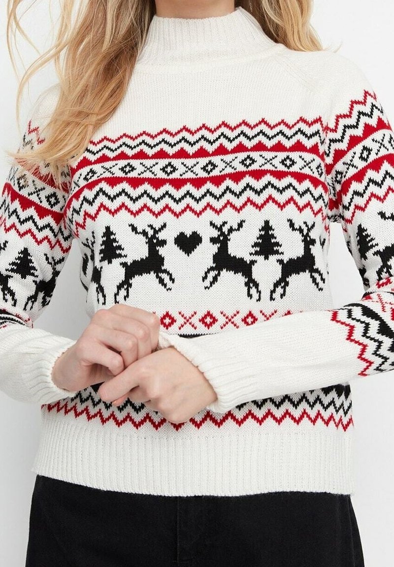 Pull en tricot blanc avec des motifs en zigzag rouge et noir, des rennes, des arbres et un cœur sur la poitrine et les manches, porté par une personne.