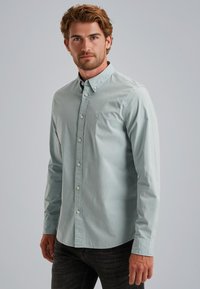 Camisa abotonada azul claro con un cuello clásico, pequeño logo en el pecho, mangas largas y dobladillo recto. El material parece suave.