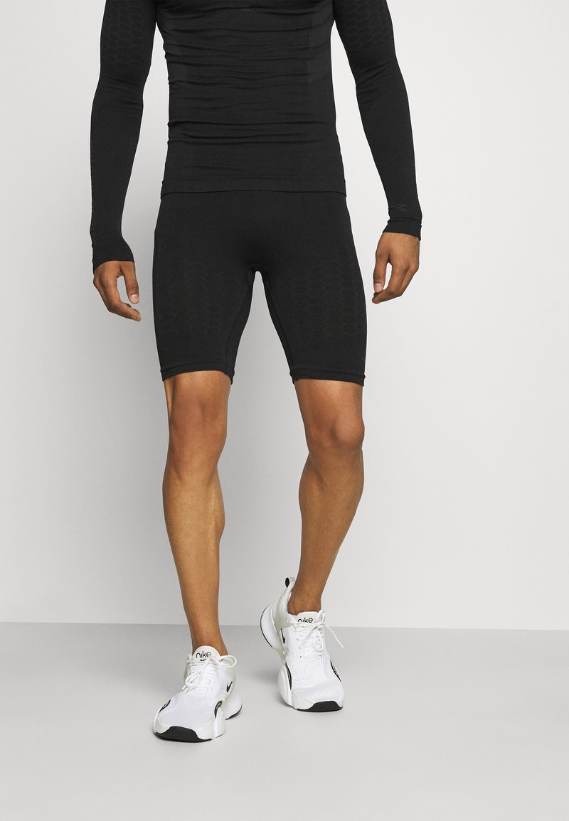 Haut de sport noir à manches longues avec un motif texturé, associé à un short noir ajusté. Chaussures de sport blanches avec des accents noirs.