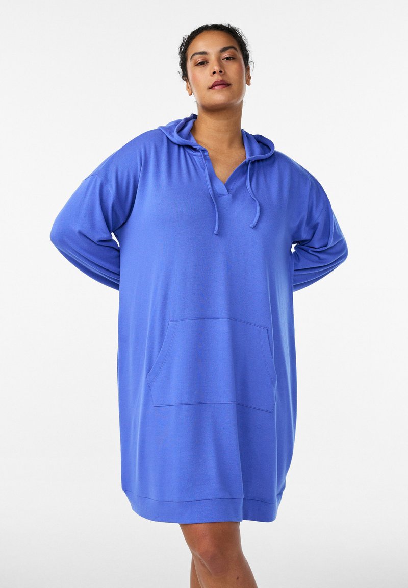 Vrouw staand met handen achter de rug, gekleed in een losse blauwe capuchonjurk met voorzak tegen een witte achtergrond.