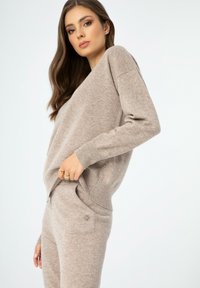 Hellbrauner Strickpullover mit lockerer Passform, weichem Material und V-Ausschnitt-Design, kombiniert mit passenden Hosen, die seitliche Taschen haben.