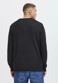 Homme avec les cheveux courts portant un pull noir à manches longues et un jeans bleu, tourné le dos sur un fond blanc uni.