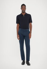 Man standing wearing a black short-sleeve polo shirt, blue trousers, and black dress shoes against a white background.