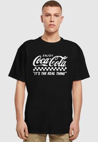Must küllane t-särk, millel on valge Coca-Cola logo kursiivis, all on ruuduline muster ja tsitaat "SEE ON TÕELINE ASI." Lühikesed varrukad, relaxed fit.