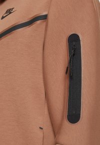 Nike fleecová mikina v světle hnědé barvě obsahuje černou kapsu na zip na rukávu, hladkou texturu, černé akcentové pruhy a detaily loga.