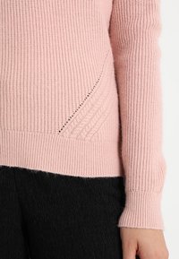 Světle růžový pletený svetr s ribovanou strukturou, který má diagonální detaily na stranách a dlouhé rukávy. Pod ním jsou viditelné tmavě černé texturované kalhoty.