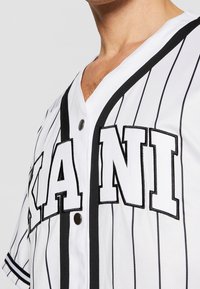 Maglia da baseball bianca con strisce nere, caratterizzata da grandi lettere in blocco "KANI" sul davanti, dettagli neri lungo il colletto e bottoni a pressione.