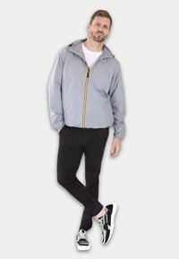 Giacca grigio chiaro con cerniera gialla e nera, polsini elastici, abbinata a pantaloni neri e sneakers nere e argento. Design semplice e funzionale.