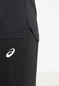 Top deportivo negro en tejido texturizado con un dobladillo trasero ligeramente más largo, combinado con leggins negros ajustados que presentan un logo blanco en el muslo.