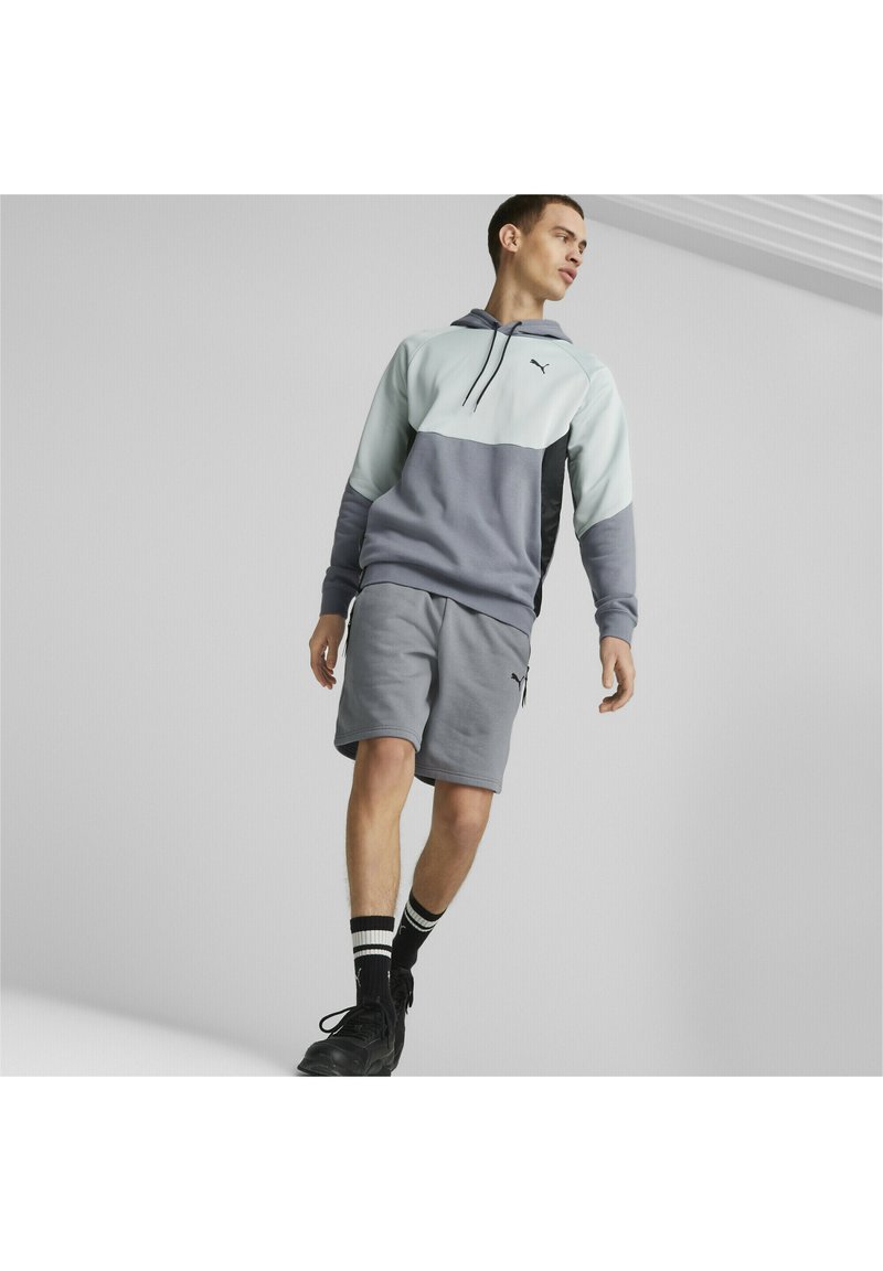 Felpa Puma in azzurro chiaro e grigio con tasca a canguro, abbinata a pantaloni corti grigi. Scarpe da ginnastica nere e calzini neri a righe completano il look.